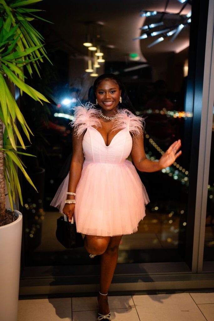 A woman in a stylish pink dress poses indoors at a nighttime event, smiling confidently.