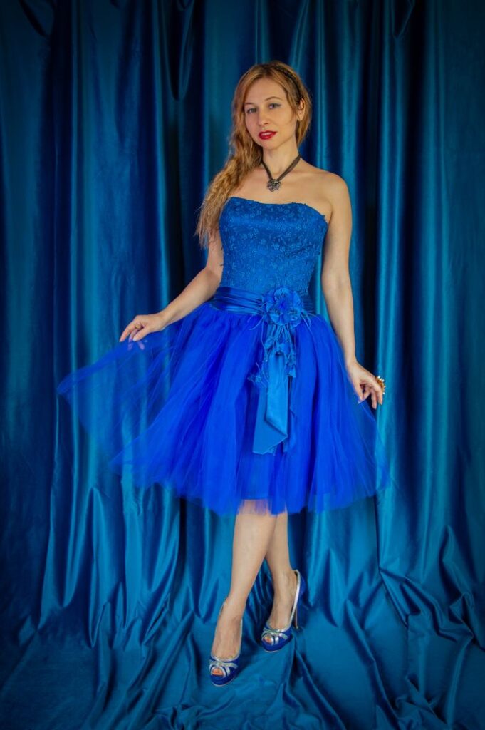 Woman elegantly posing in a strapless blue dress with a draped fabric background.