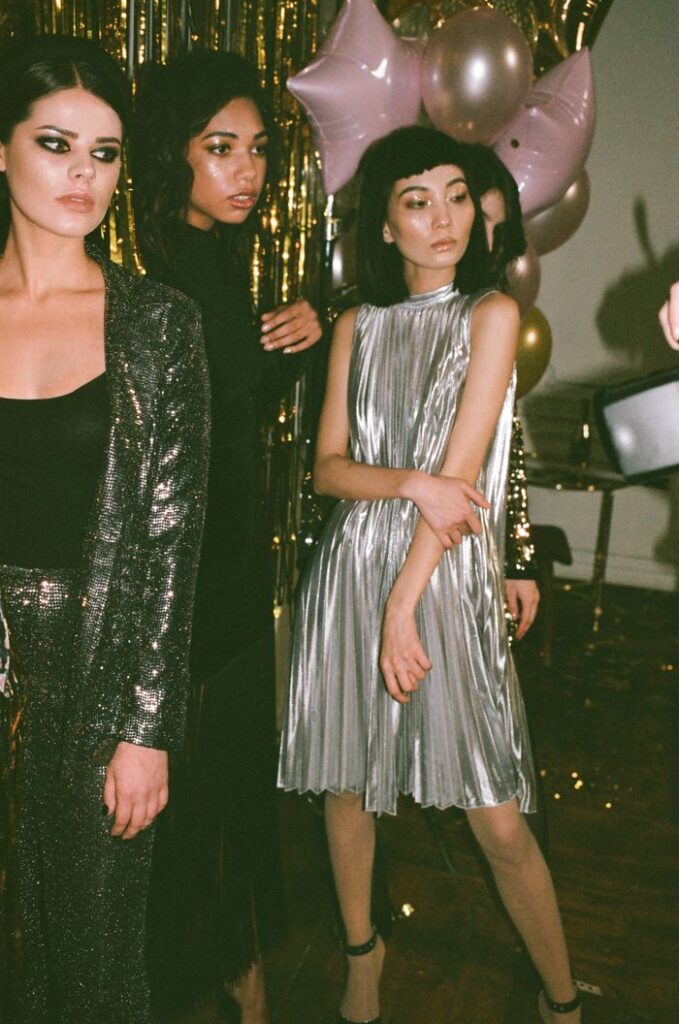 Crop young women in shiny wear with makeup standing on floor near assorted balloons during festive occasion